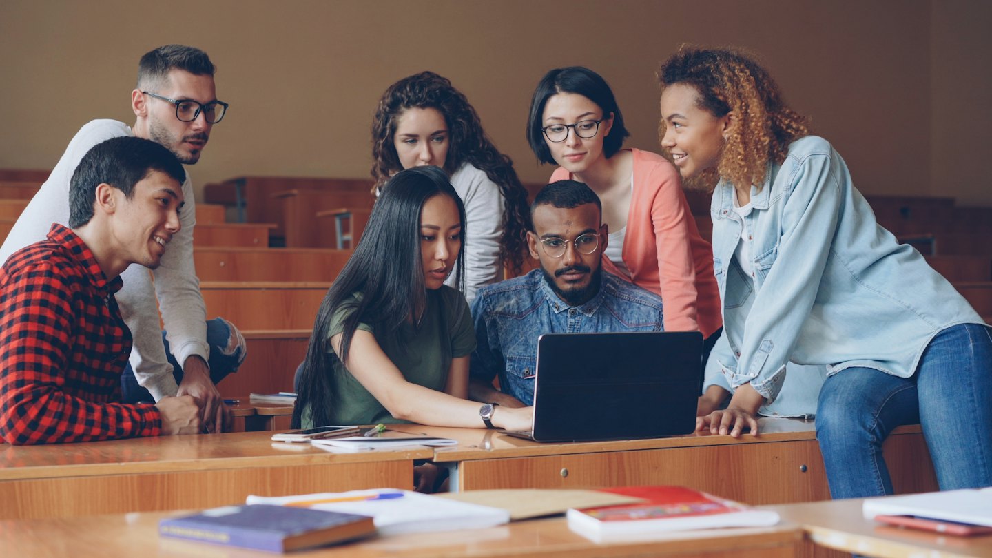 University students collaborating around a laptop in a lecture hall.